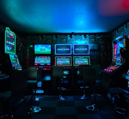 Dimly lit gaming arcade featuring a variety of brightly illuminated slot machines and video gaming screens. Neon signs and colorful displays create a vibrant atmosphere. The arrangement of gaming machines suggests an inviting space for entertainment, with chairs positioned for players.