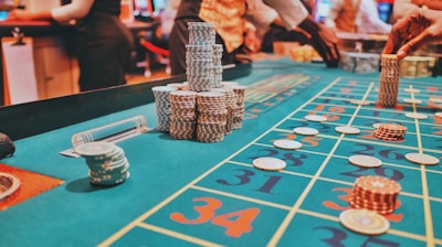A casino scene with a roulette table features prominently, covered in green felt with colorful numbers and betting squares. Several stacks of poker chips are placed on different numbers, and the hands of several people are visible as they interact with the table. In the background, there are slot machines and more people, creating a vibrant and busy atmosphere.