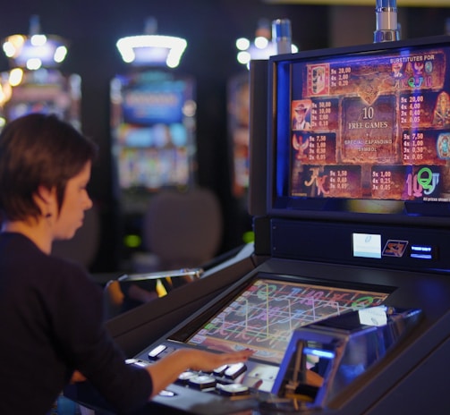 A person is seated at a slot machine console in a casino, focusing intently on the game displayed on the screen. The background shows several other illuminated gaming machines, adding a vibrant and colorful atmosphere to the scene.