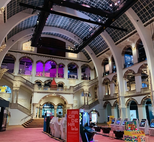 The interior of a large, ornately designed building with arched ceilings and intricate architectural details. The space features gaming machines lined up along the floor, under a high ceiling with a grid pattern. Lighting in purple and white tones enhances the ambiance.