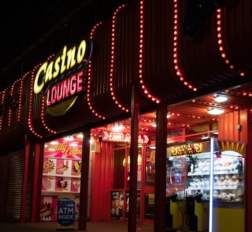 A brightly lit casino lounge entrance adorned with neon lights. The exterior features colorful signage including the words 'Casino' and 'Lounge' in vivid colors, with additional lights framing the building. Inside, a glimpse of a claw machine filled with plush toys and arcade games is visible. The area exudes an inviting yet bustling atmosphere typical of entertainment venues.