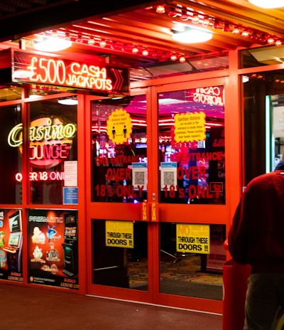 A brightly lit entrance to a casino with neon signs, including one advertising a £500 jackpot. The doors have clear signage indicating entry is restricted to those over 18. A person in a red jacket stands to the right, possibly interacting with a machine or display.