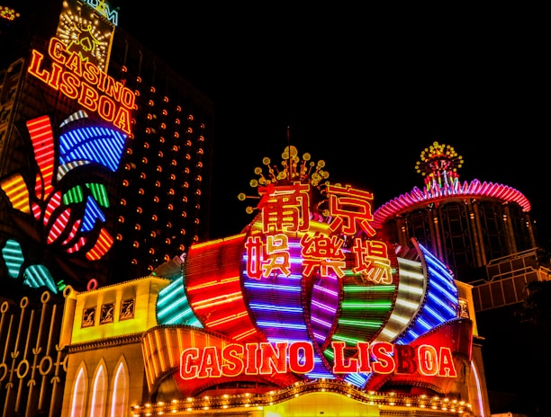 Bright neon lights and colorful signage adorn the exterior of Casino Lisboa, creating a vibrant and lively atmosphere. The façade features bold, multicolored designs with a mix of English and Chinese characters prominently displayed. The lights form intricate patterns and shapes, contributing to the energetic ambiance of the scene.
