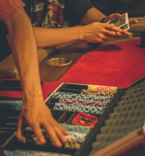 A person reaches into a poker chip case on a red felt table, while another person holds playing cards. The scene is dimly lit, creating a warm and engaged atmosphere.
