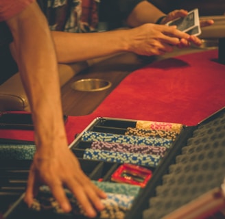 A person reaches into a poker chip case on a red felt table, while another person holds playing cards. The scene is dimly lit, creating a warm and engaged atmosphere.