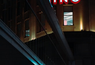 A night scene featuring a building with illuminated neon signage displaying 'CASINO'. The environment is urban with reflections on glass windows and glowing street traffic lights below. The atmosphere is dimly lit, with artificial lighting casting vibrant colors.