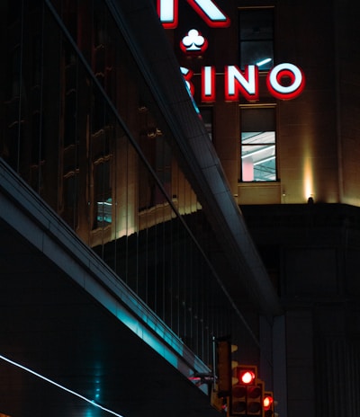 A night scene featuring a building with illuminated neon signage displaying 'CASINO'. The environment is urban with reflections on glass windows and glowing street traffic lights below. The atmosphere is dimly lit, with artificial lighting casting vibrant colors.