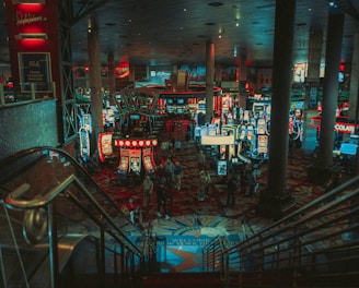 A bustling casino floor filled with groups of slot machines and people walking around. The atmosphere is vibrant with neon lights and illuminated signs. The interior has a ceiling supported by large pillars and there are stairs leading down to the gaming area.