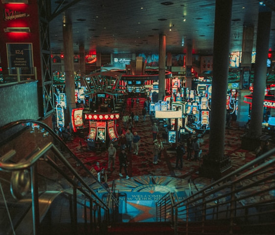 A bustling casino floor filled with groups of slot machines and people walking around. The atmosphere is vibrant with neon lights and illuminated signs. The interior has a ceiling supported by large pillars and there are stairs leading down to the gaming area.